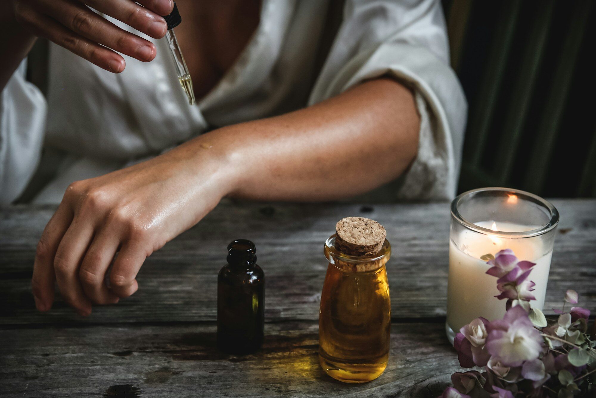 Hands using a dropper of botanical facial oil beside amber bottles, a lit candle and orchid stems on weathered wood